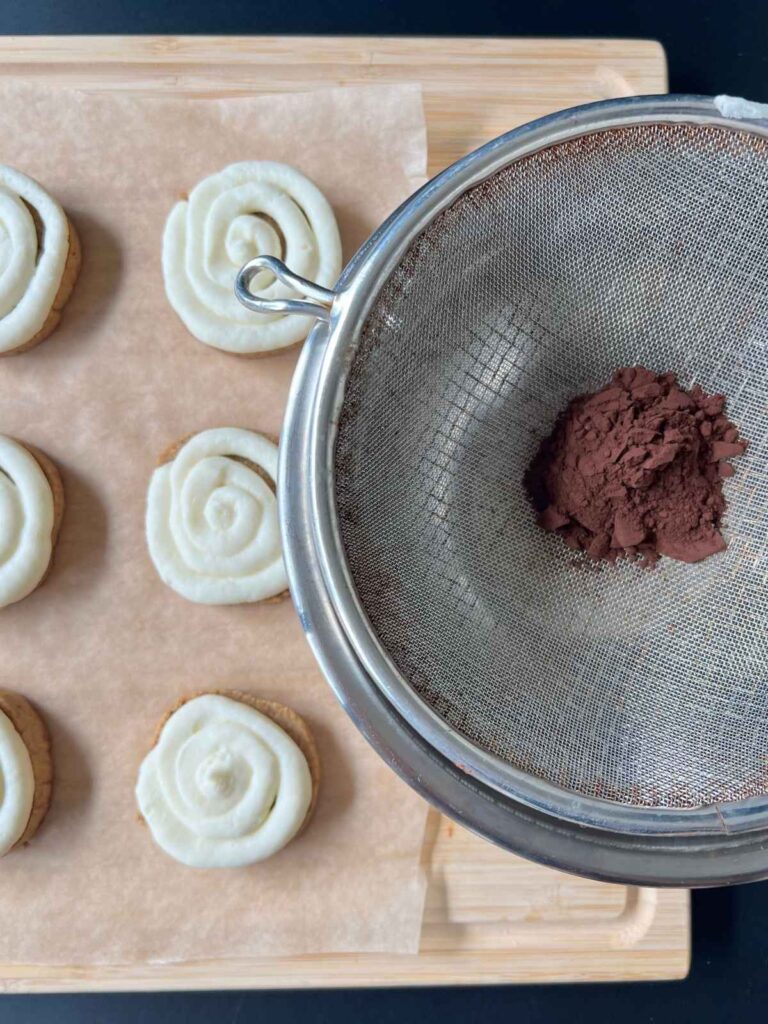 cocoa powder in a fine mesh strainer above the frosted cookies